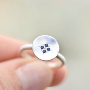 Silver ring with flat silver disc on face, set with four blue sapphires. The ring is held aloft.