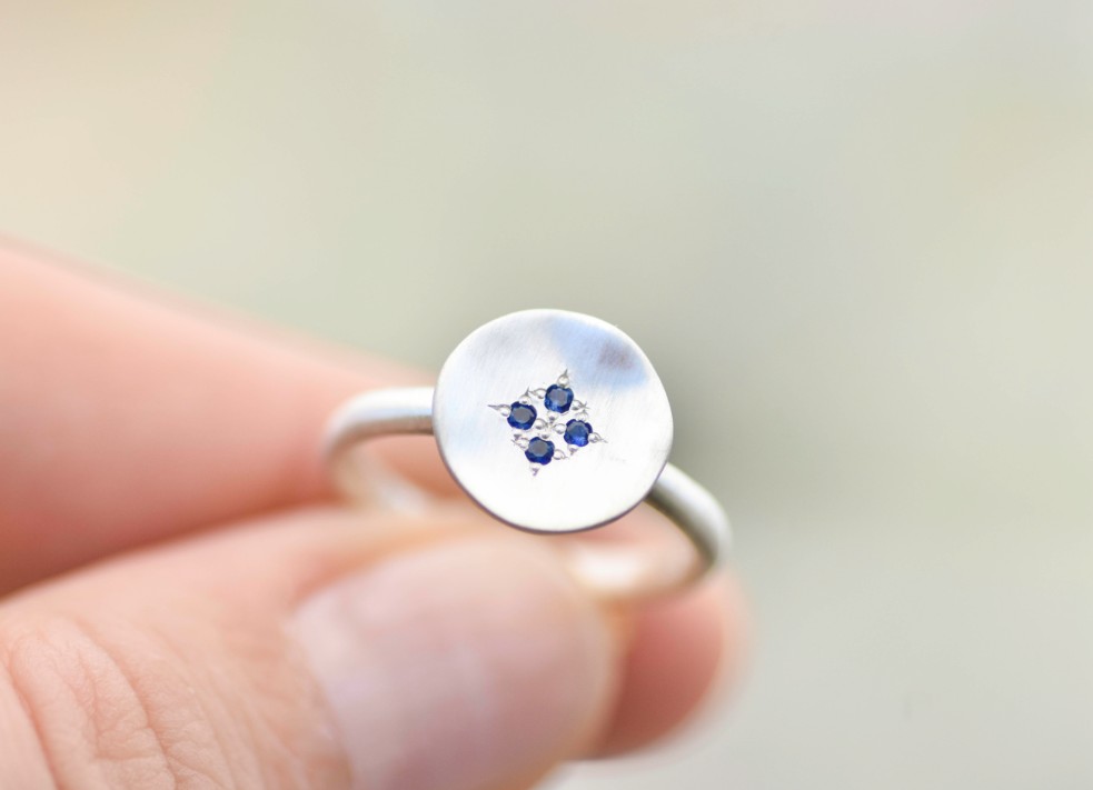 Silver ring with flat silver disc on face, set with four blue sapphires. The ring is held aloft.