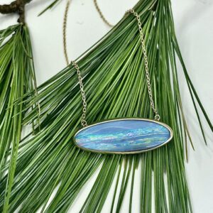 Boulder opal slab, bezel-set in gold, suspended on a two points of a necklace chain. The jewelry is laid on pine needles.
