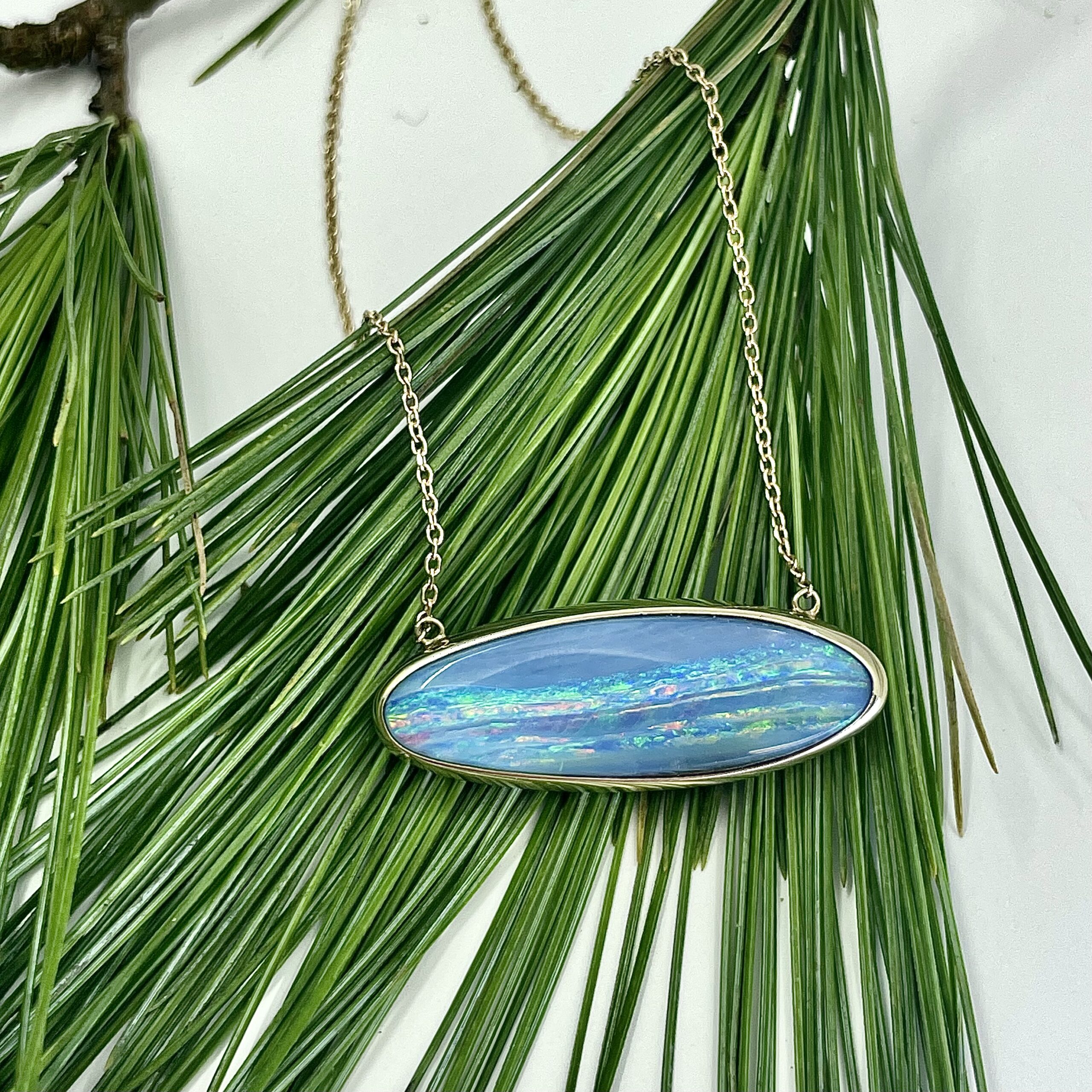 Boulder opal slab, bezel-set in gold, suspended on a two points of a necklace chain. The jewelry is laid on pine needles.